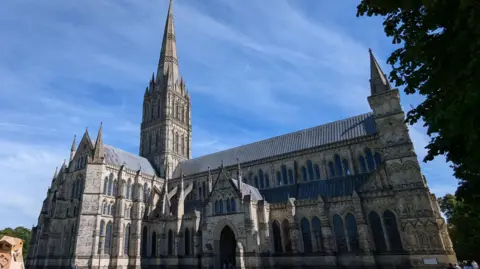 Salisbury Cathedral under a bright blue sky - a grand medieval building with a tall spire and lots of arched windows