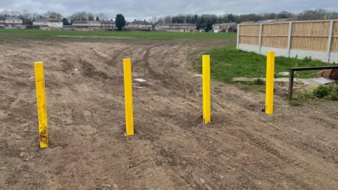 Simon Thake Four yellow metal barriers stand vertical in a dirt path leading to a grass field. A row of houses backing onto the field can be seen in the distance.