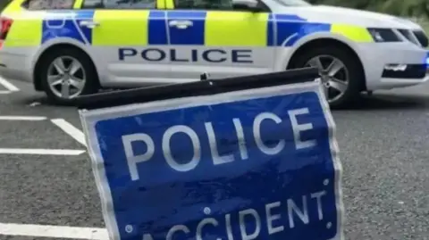 A police car parked on a road with a blue sign which reads 'police accident' positioned in the foreground.
