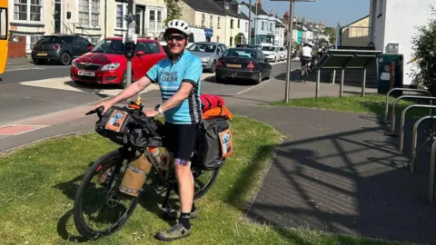 Norman Brown A man with a bike stands on the grass. His bike is loaded up with supplies in panniers. Behind him is a street with cars and a row of houses. He is wearing a cycle helmet, sunglasses and blue and black cycling gear,
