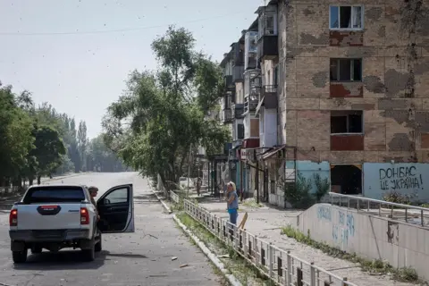 Alina Smutko/Reuters A resident stands on a street outside an apartment block, communicating with a police officer who is poking their head out of a police van, in Toretsk near the frontline in Donetsk region in July 2024. 