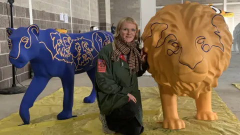 A woman with short blonde hair and glasses is wearing an outdoor coat and scarf and crouching on plastic sheeting. She is next to a bright blue lioness sculpture and a bright yellow lion sculpture