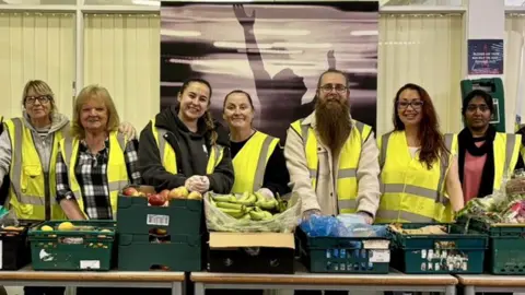 Some of the volunteers at Helping Hearts, all wearing yellow hi-vis vests, stand behind a row of tables containing green plastic baskets full of food