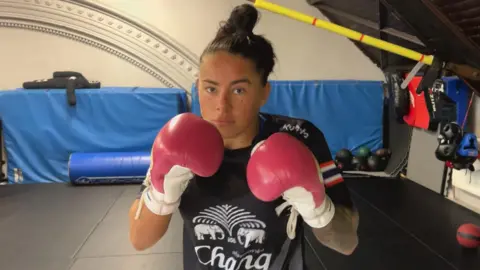 Kennedy Freeman wearing pink and white boxing gloves and standing in a boxing pose, with a black t-shirt with white elephants on it, hair tied up in a bun, with gym equipment in the background.