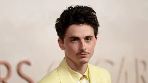 Getty Images Timothée Chalamet wears a butter yellow suit with a gold pin on the collar. 