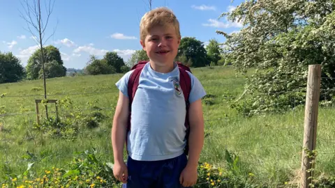 Macaulay is standing in a flowering field and smiling widely at the camera. He has short blond hair and is wearing a blue top and shorts.