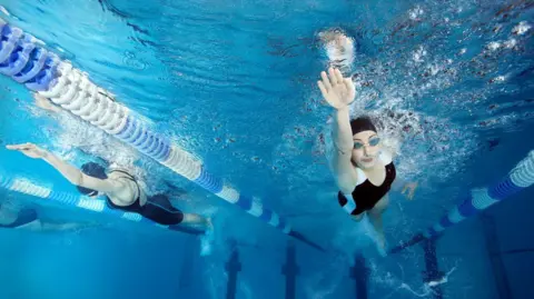 Two women in black swimming costumes and caps swimming in a pool, seen from below