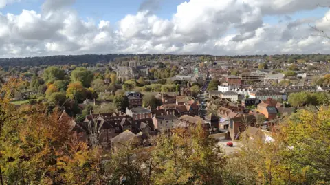 An aerial view over the centre of Winchester. In the centre of the frame you can see the large stone cathedral. The city is made up of stone and brick buildings and surrounded by trees that are turning shades of orange and brown.