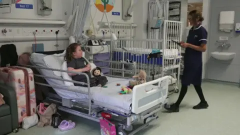 A nurse approaches a young girl sat in her hospital bed. She has cuddly toys on her bed.