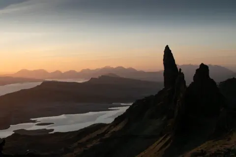 Kate Masson The Old Man of Storr - a sharp rock formation on a dark hillside - at sunrise. It overlooks calm water and distant misty mountains. The sky has an orange tinge.
