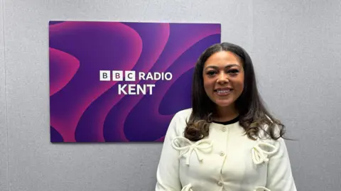 An image of Tia Davies stood in front of the BBC Radio Kent logo. Tia is wearing a white jacket with bows. 