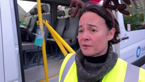 Lesley Leeson is standing in front of a minibus. She is wearing a grey jumper and a high-vis jacket. 