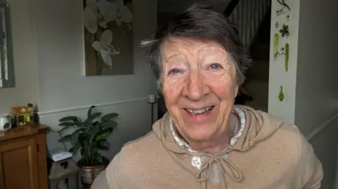 Robby West/BBC A woman wearing a beige jumper stands in her hallway. The walls are pale with pictures and a plant can be seen in a pot. 