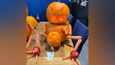 A carved pumpkin "head" sits on another slightly bigger pumpkin "body", and appears to be giving birth to a smaller carved pumpkin. Carrots make up the arms and legs. Each foot is one red apple. The creation, which has two candles in its mouth, is sitting on a wooden dining table.