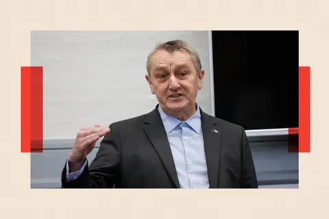 EPA Wales Green Party Leader, Anthony Slaughter. He is wearing a dark suit and a blue shirt buttoned up at the collar. He raises his right hand as if for emphasis. Behind him a white wall and a blackboard are visible.