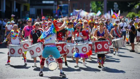 PA Media A shot looking down a large pride parade - in the foreground are a samba band, all holding red and white drums and dressed for a summer's day with rainbow accessories. The leader has his back to the camera to face and instruct the band, and wears a pair of rainbow wings