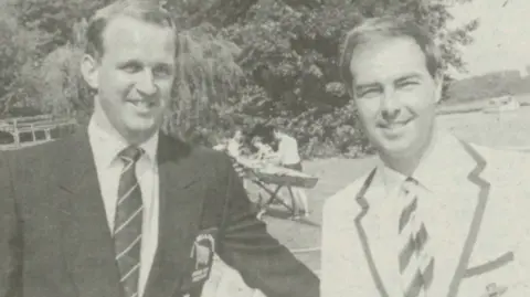Henley Standard A black and white photo of two men dressed in blazers with striped ties grinning at the camera. Behind them is a river with several people working on rowing boats. 