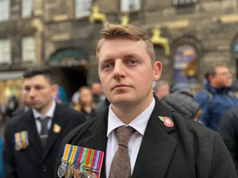 A young man with short sandy hair, wearing a black overcoat adorned with a red poppy brooch and a row of medals.