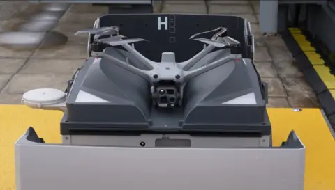 A grey four-rotor drone sitting inside an open docking and charging box.