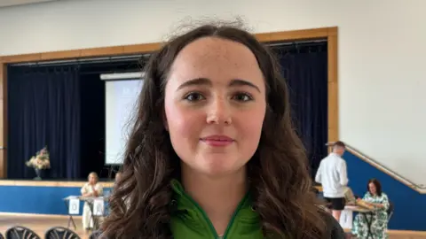 Helena smiling in front of the camera. She has long curly brown hair. There are school chairs behind her in an assembly hall with teachers in the background.