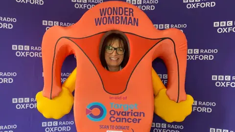 Lis Hale has medium length dark hair and glasses. She is wearing a costume that looks like a pair of ovaries, and has 'Target Ovarian Cancer' written on the front. Behind her is a purple wall with 'BBC Radio Oxford' on it in white writing.
