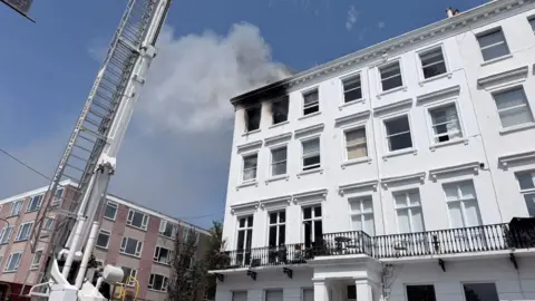 Smoke coming out of a room in a six-storey building. There is an aerial ladder from the fire service dealing with the incident. 