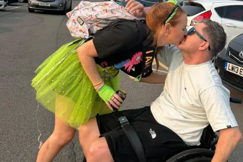 Anna Thomas A brown-haired man wearing a white T-shirt, black shorts, and sunglasses was sitting in a wheelchair in a parking lot with cars in the background. A red-haired woman wearing a black T-shirt and a neon green tutu bends over and kisses him.