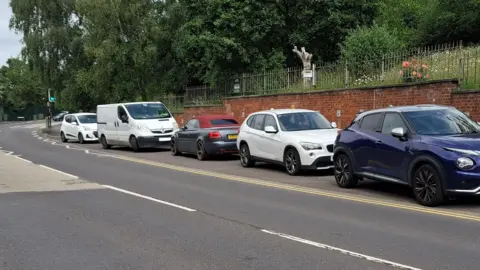 Cars and a van of various colours parked on a layby just past the bend of a road. Behind the cars is a red-brick wall topped with iron railings. Behind the railings is a grassy bank topped with green trees.