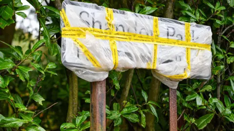 Pacemaker A sign wrapped in plastic sits in front of green foliage. It's wrapped in yellow tape. The two legs of the sign are rusted. There are brown leaves on the ground. 