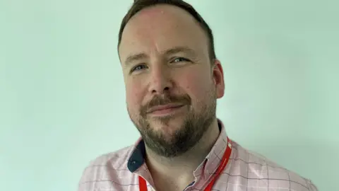 RCN Simon Browes, who is wearing a pink checked shirt with a red lanyard around his neck. He has a beard and brown hair and is standing in front of a pale green background.