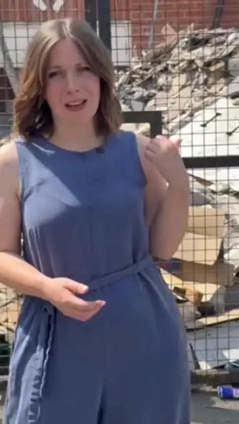 A woman wearing a loose blue jumpsuit stands in front of a large pile of waste behind a fence. She has a concerned expression and is pointing at the mess behind her. 