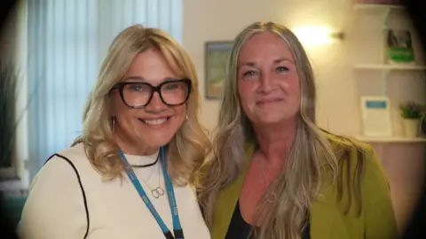 Jamie Niblock/ BBC Louise is wearing a white top, with a blue lanyard, and glasses. She is smiling, standing next to Julie, who is wearing a green suit jacket, and a black top. She is also blonde, and smiling. They are photographed in the main reception of the Macmillan information centre at West Suffolk Hospital