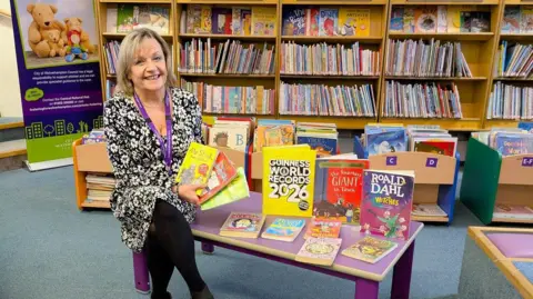Handout Elisabeth Whitehouse sits on a table in a library children's section. She is holding all the favourite books of the moment and sitting next to a copy of the Guinness World Records 2026 and novels by Roald Dahl.