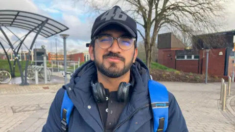Lingadhar wears a dark winter coat, a blue backpack, a black cap and a pair of headphones around his neck. He stands outdoors near a bike shelter. 