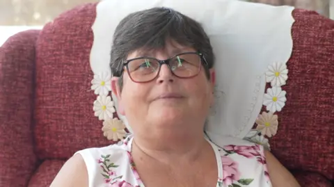 A woman is sitting in a pink armchair. She is wearing a white dress with large pink flowers on it and glasses. She is looking into the camera and smiling.
