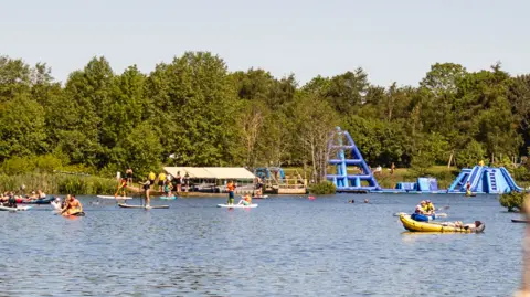 Alderford Lake A lake surrounded by trees with people in boats and some large blue inflatable slides in the background