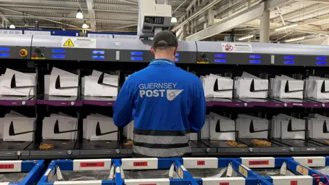 BBC Photo of a man's back in a blue jacket with 'Guernsey Post' written on the back. He is standing in front of a large letter sorting grid.