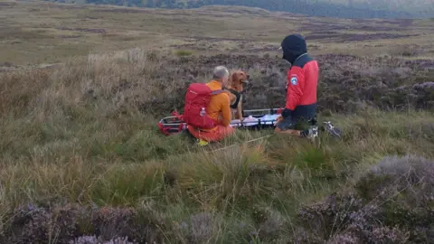 Kendal MSRT Two mountain rescue volunteers sit with a dog on a stretcher across long grass. The volunteers are wearing bright orange and red clothing. The dog is sat upright and is being treated for its injury.
