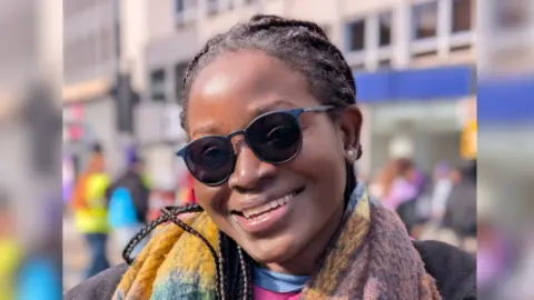 Favour has brown hair and is wearing a multi-coloured scarf and black sunglasses.