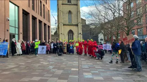 A group of protestors, some wearing red robes, pose with banners outside a Medieval church next to a red panelled building.   