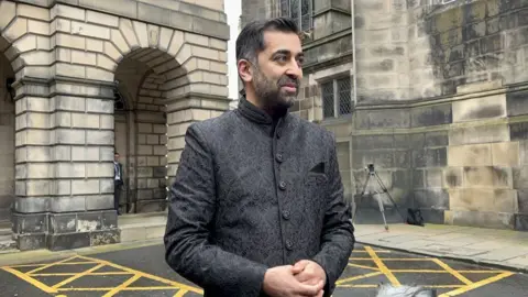 Getty Images Humza Yousaf standing in Parliament Square, Edinburgh. He is wearing a dark, buttoned-up traditional shalwar kameez and is smiling.