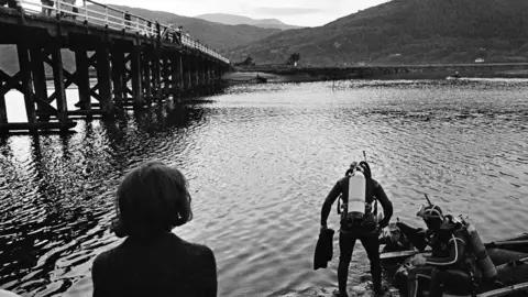 Mirrorpix via Getty Images Black and white image looking across a stretch of water, with a large wooden bridge on the left crossing an estuary. In the foreground, a woman with her back to the camera is looking out, as two divers in scuba gear prepare to enter the water. One is standing with flippers in his hand and a white air tank on his back - the other is sat in a boat on the water's edge