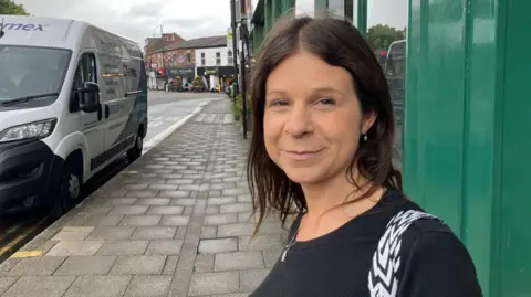 LDRS Lynsey Lowdon is standing on the high street in Cheadle. In the background is a number of shops. She is wearing a black top and has a black and white bag over her shoulder.