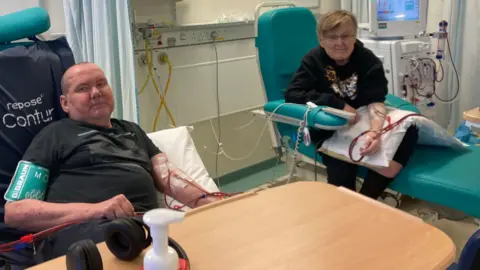 The image shows a balding man in a black t-shirt on the left of the image, next to a man with blond hair wearing a black hoodie. Both are undergoing renal dialysis at Coventry's University Hospital. 