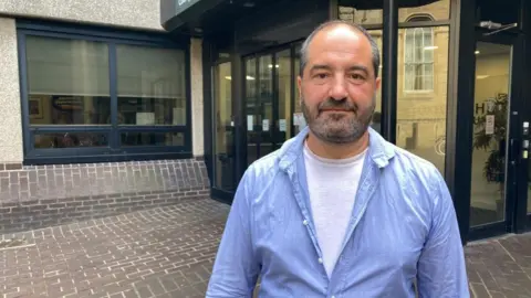 A man is standing outside a council office. He is wearing a white t-shirt, with a blue shirt over the top. He has short hair and a beard. 