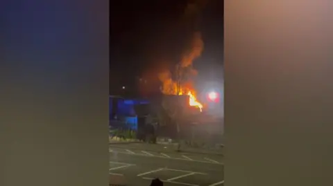 A screenshot of the fire filmed on a phone. The filming is across a road and there is a blue metal building in front of a blaze of fire at night time. 