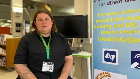 Steph looks at the camera with the library behind, to her right is a banner on Deaf Awareness Week