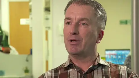 A man wearing a brown patterned shirt is talking to the camera from his work office. He has short grey hair.