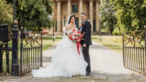 A woman in a white wedding dress stands with a man in a suit. She holds a bouquet of red and pink flowers with a red ribbon. They stand at the pathway to a round church, metal ornate gates are open next to them and the path lined with traditional street lights. To the right and left is a lawn with large trees.