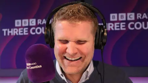 JVS sitting in the studio behind the microphone, wearing black headphones over his ears. He is chuckling. The purple mic says BBC Three Counties, which is also written on studio walls behind him. He has short blonde hair and salt and pepper coloured stubble. He looks towards the mic but not directly at the camera.
